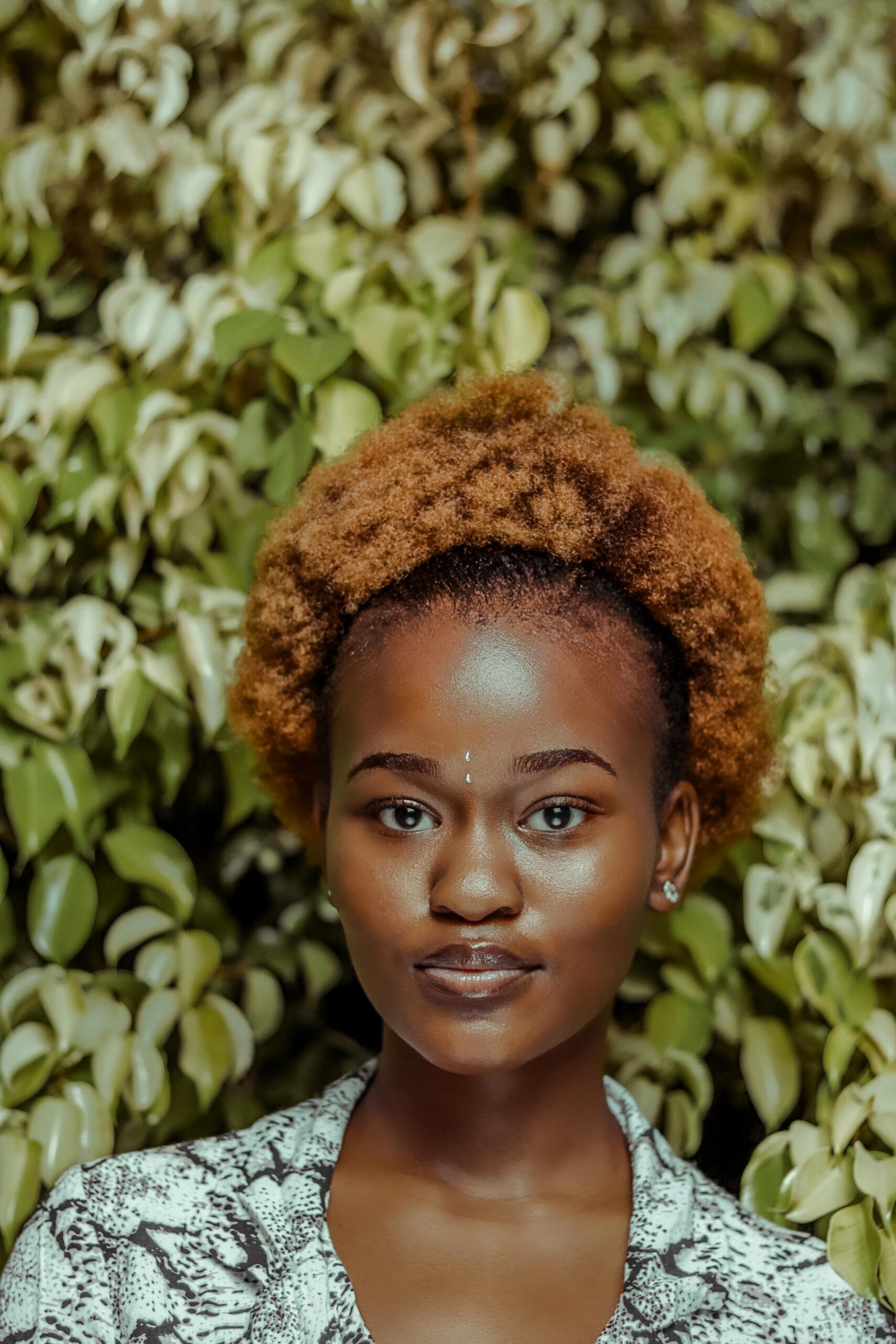 Portrait of a young woman with an afro hairstyle in front of vibrant green foliage.